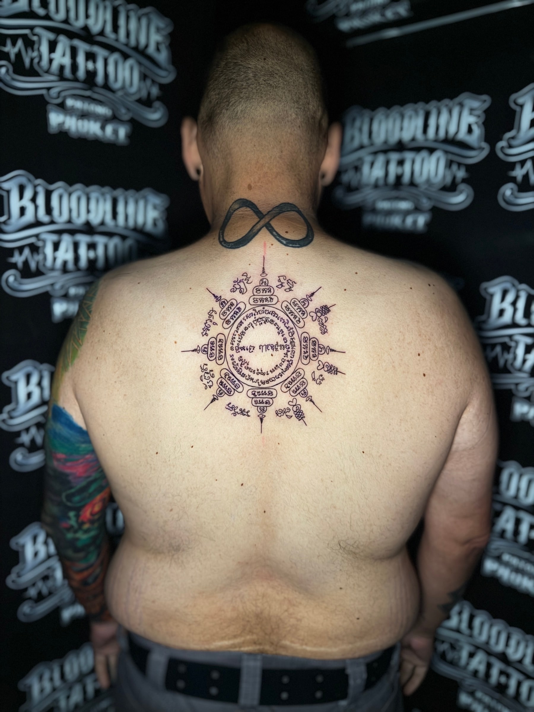 Paed Tidt (Eight-Directional Yant) Sak Yant with concentric Khom script and eight radiating unalome spires on the upper back, with a solid infinity symbol on the nape at Bloodline Tattoo Phuket.