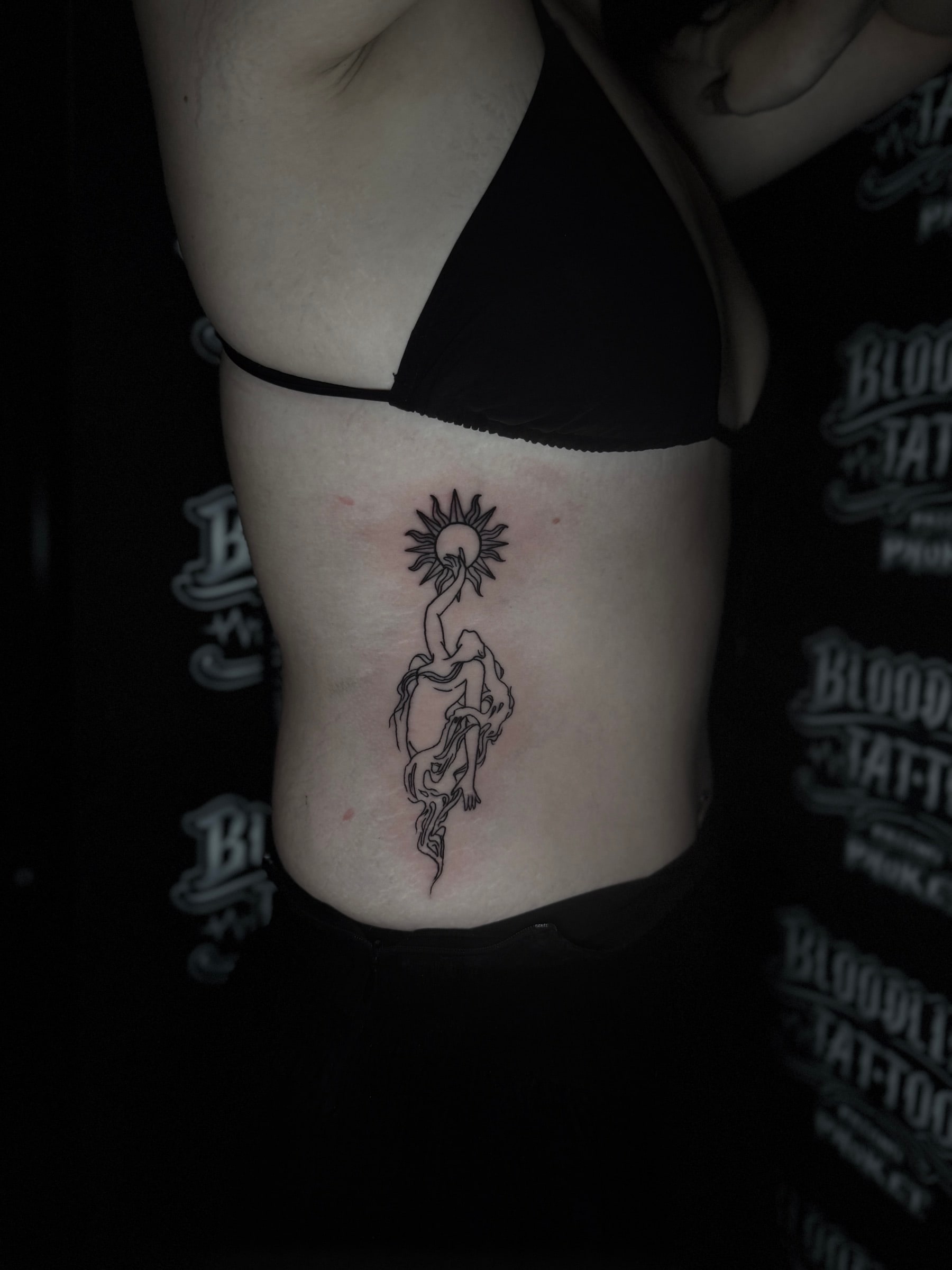 Fine-line bamboo tattoo of a stylized woman reaching toward a radiant sun on the side ribs at Bloodline Tattoo Phuket.