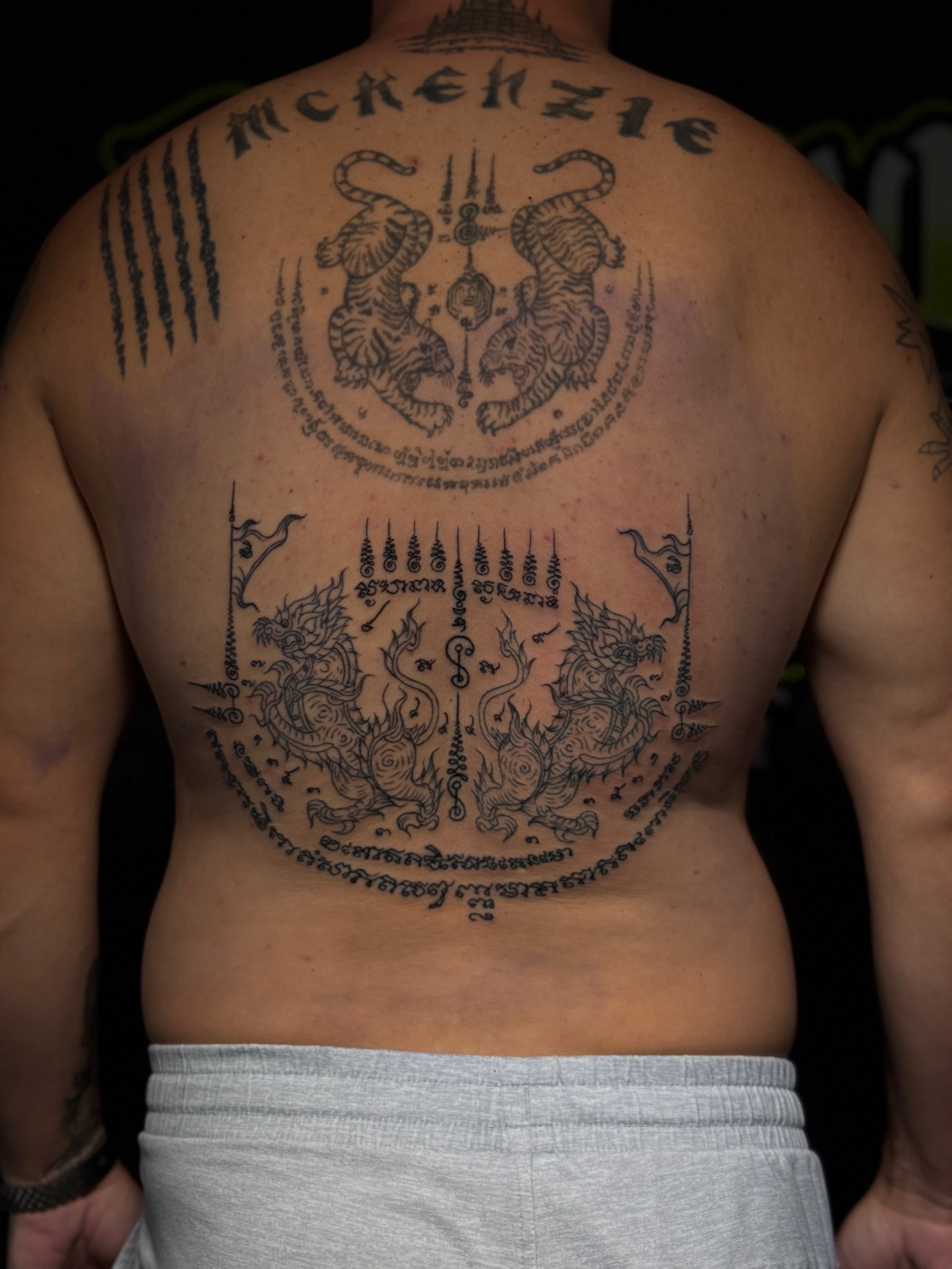 Large back Sak Yant with Suea Koo twin tigers, Yant Singha mythical lions, Hah Taew five lines, and MCKENZIE script lettering at Bloodline Tattoo Phuket.