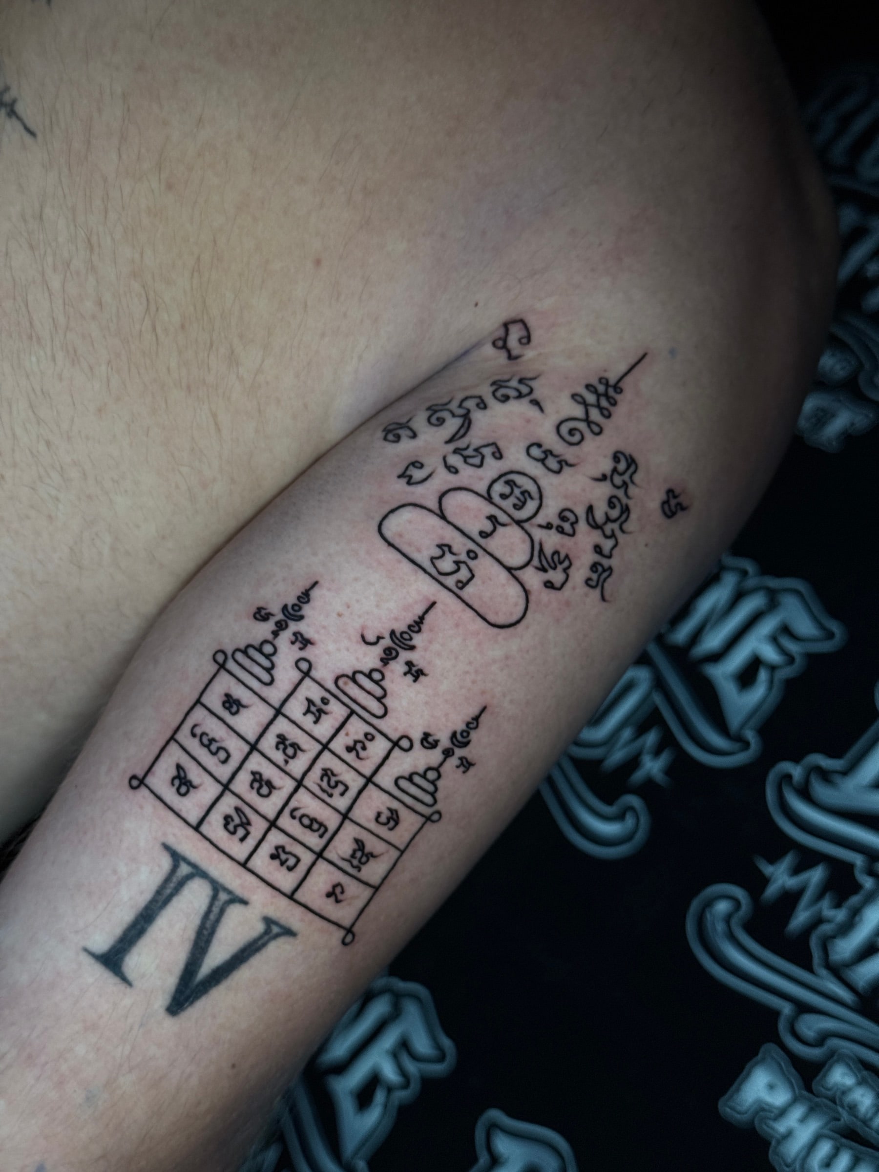 Sak Yant talismanic grid yantra with sixteen Khom script cells, tiered script design, unalomes, and Roman numeral IV on the inner arm at Bloodline Tattoo Phuket.