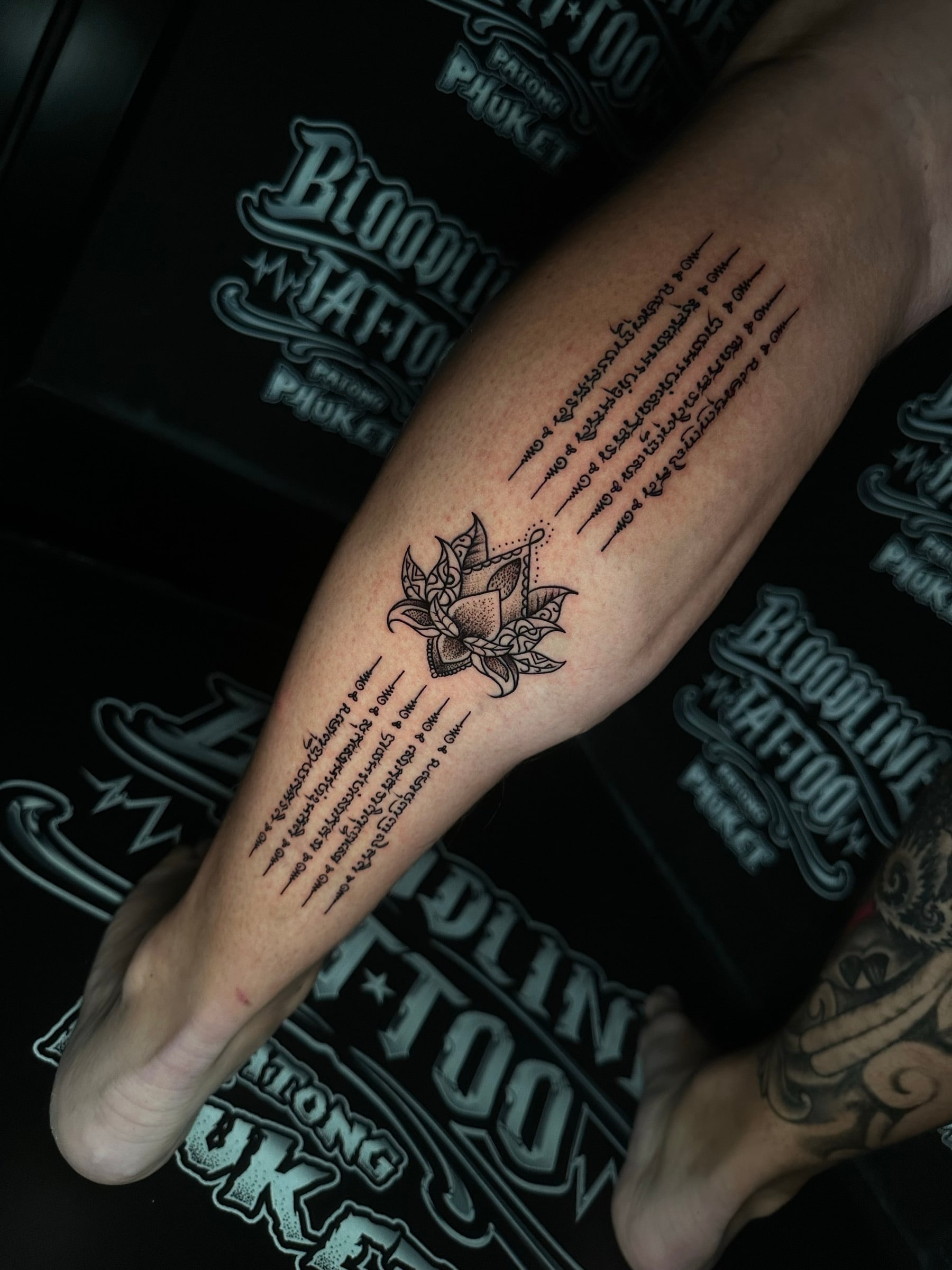 Hah Taew-style Sak Yant with a decorative lotus motif and five rows of Khom script above and below on the lower leg at Bloodline Tattoo Phuket.