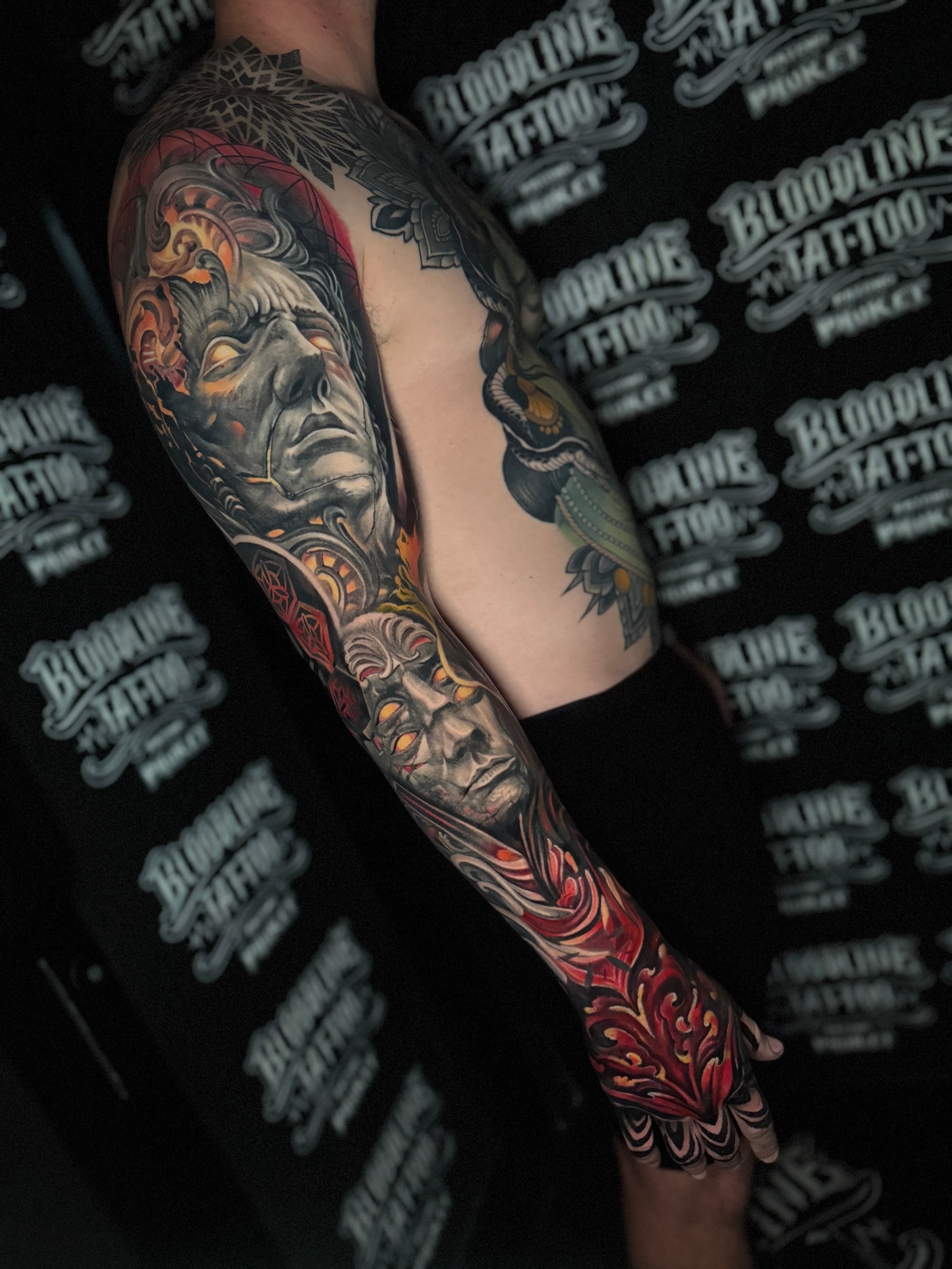 Colour sleeve and hand tattoo with realistic grey portraits, glowing yellow eyes, red ornamental patterns, and a shoulder mandala at Bloodline Tattoo Phuket.