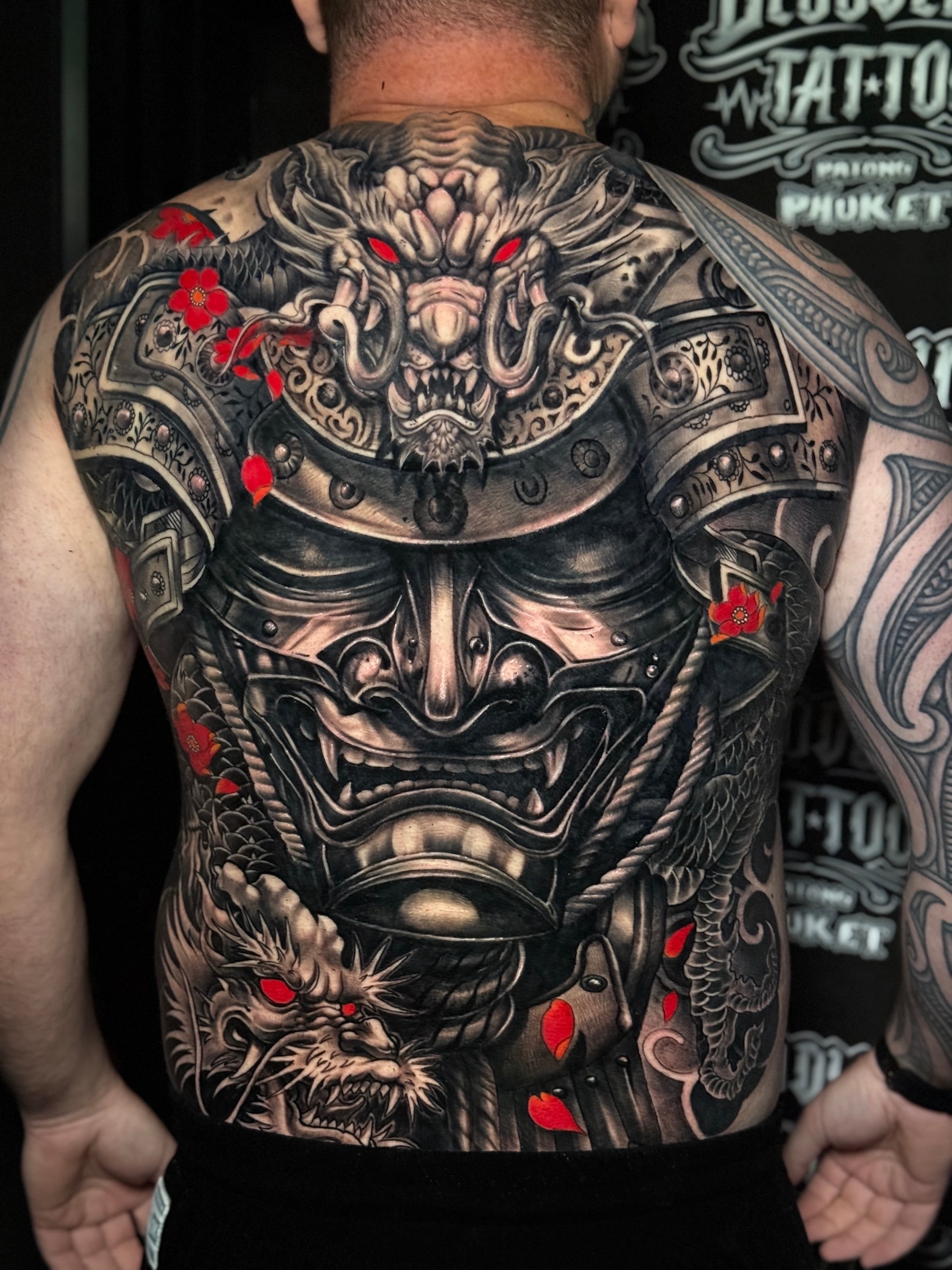 Japanese black and grey full-back tattoo with samurai menpo and kabuto, flanking dragon heads with red eyes, and red cherry blossoms at Bloodline Tattoo Phuket.