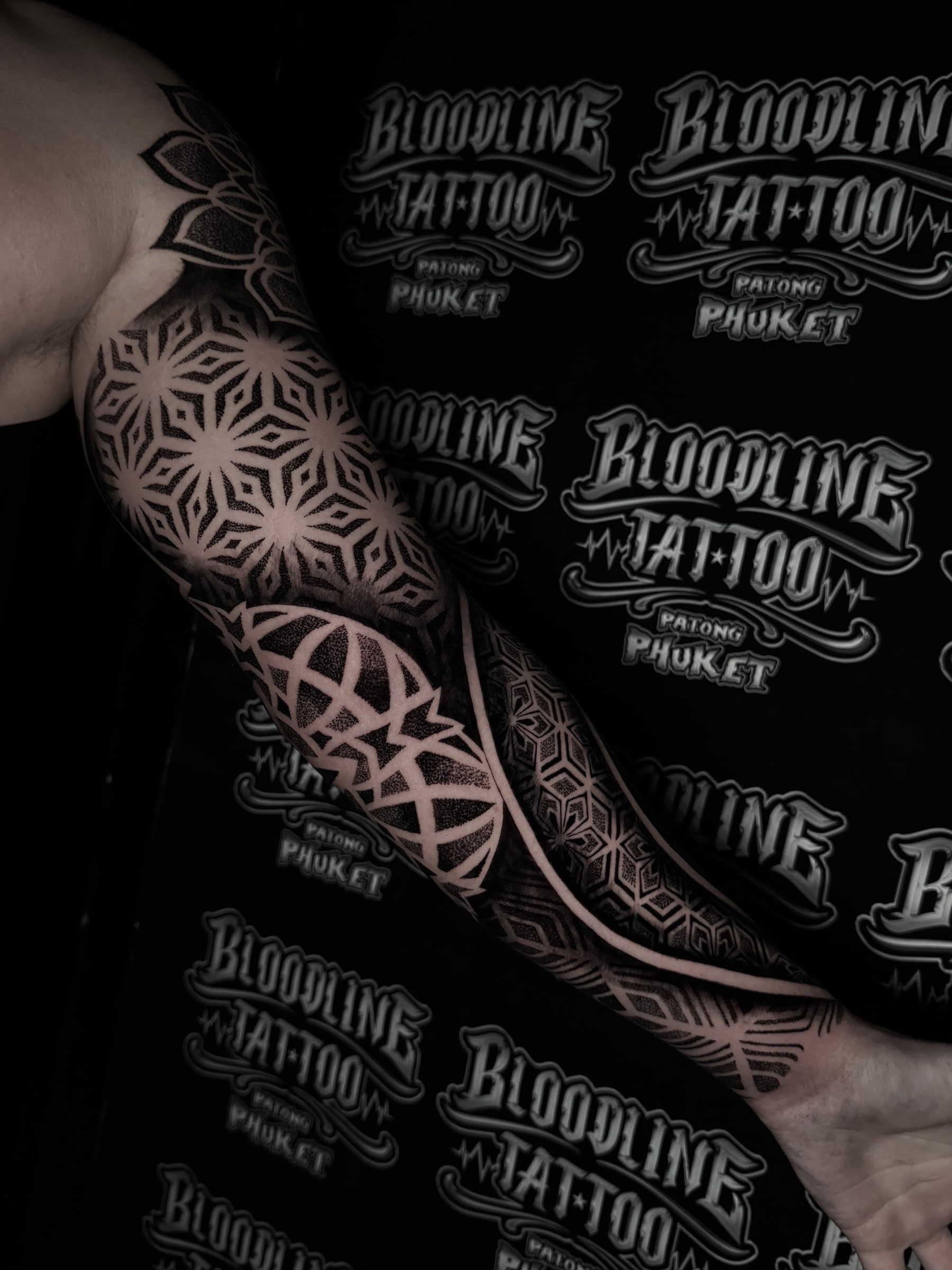 Geometric mandala sleeve with star-shaped upper arm mandalas, a sweeping negative-space curve, and stippled hexagonal wrist fill at Bloodline Tattoo Phuket.