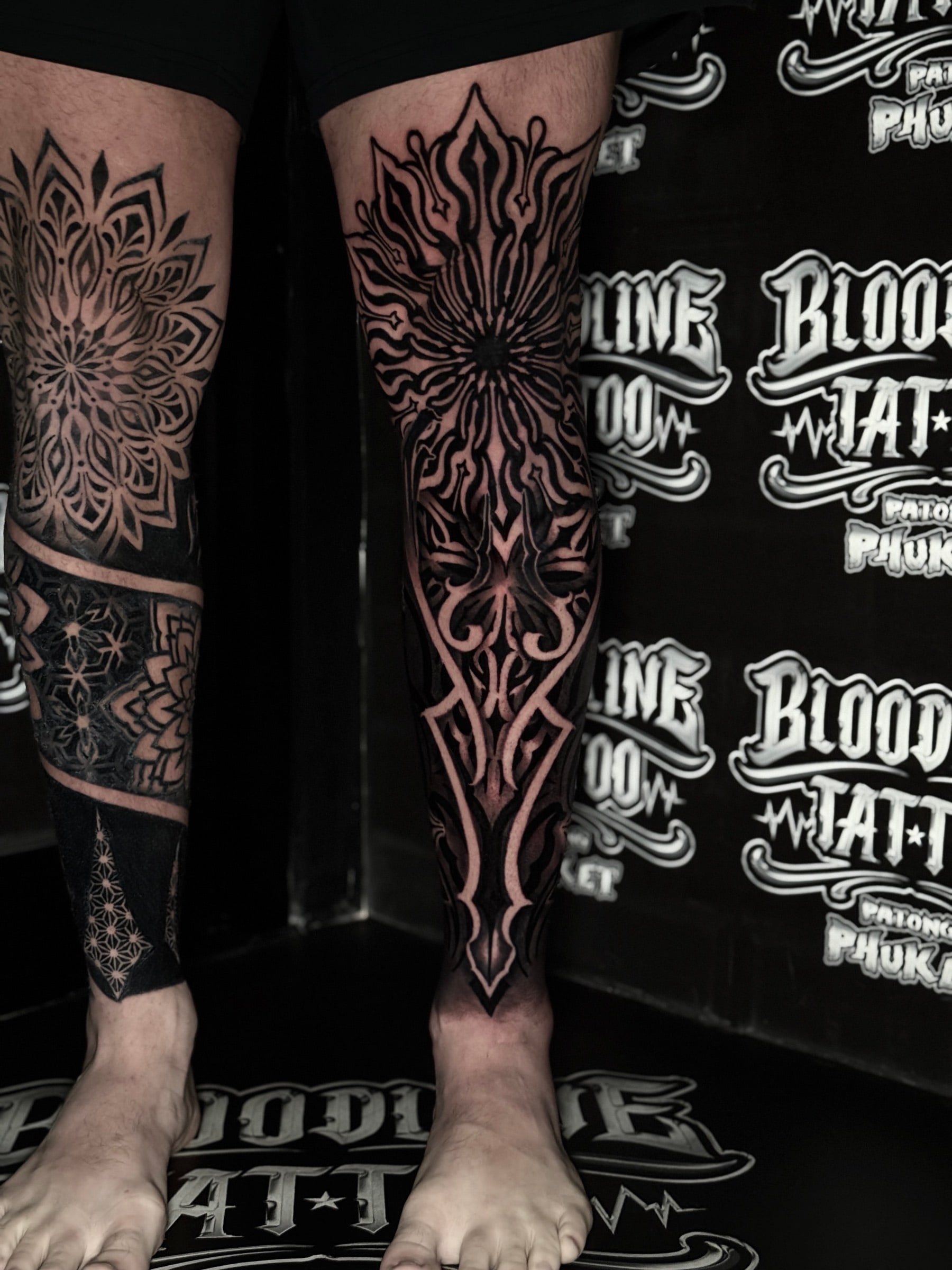 Dual-leg mandala blackwork with radiating knee mandalas, interlocking star shin bands, and bold symmetrical lower-leg geometry at Bloodline Tattoo Phuket.